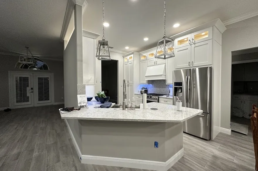 White kitchen remodel with a large angled quartz island, lantern pendants and lit glass-front cabinets by Posada Remodeling & Design